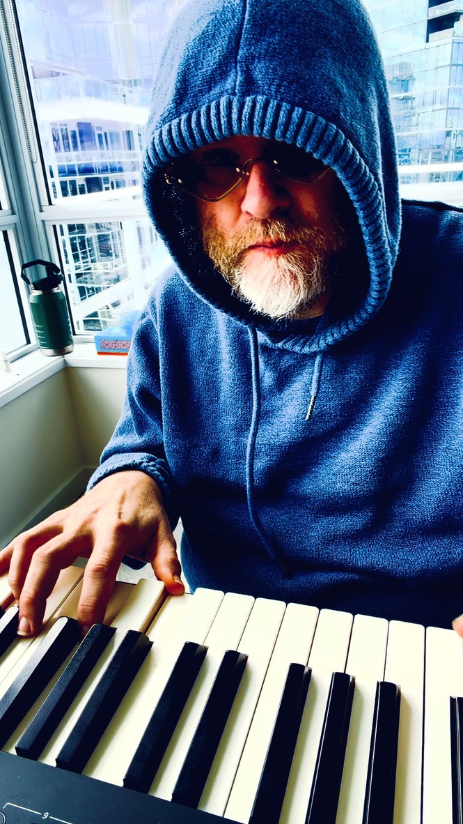 A hooded musician playing an electronic piano beside tall windows.