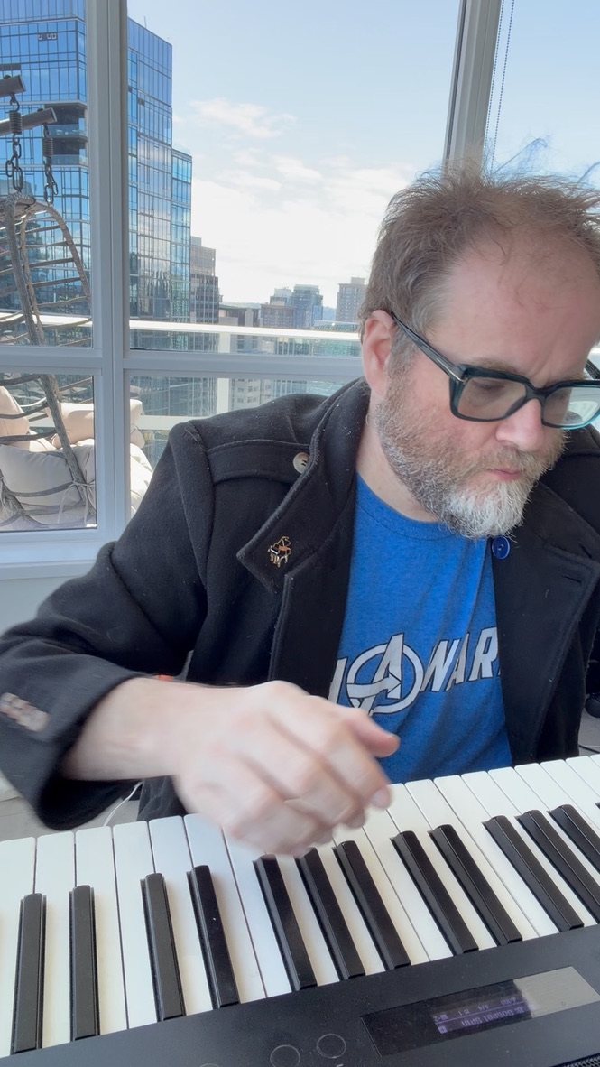 A musician in glasses striking keys on an electronic piano in a bright room.